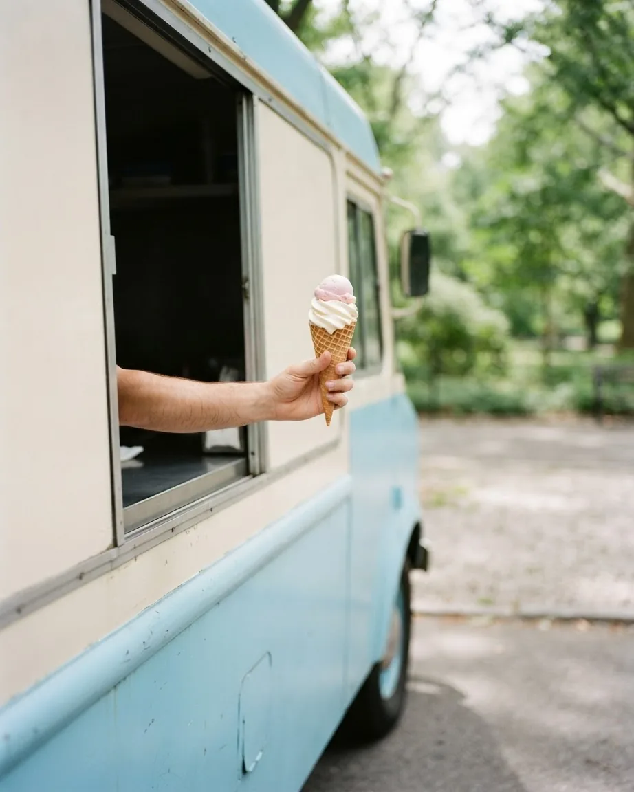 A forearm extends from the open service window of a pale blue ice cream truck, holding a two-scoop ice cream cone.